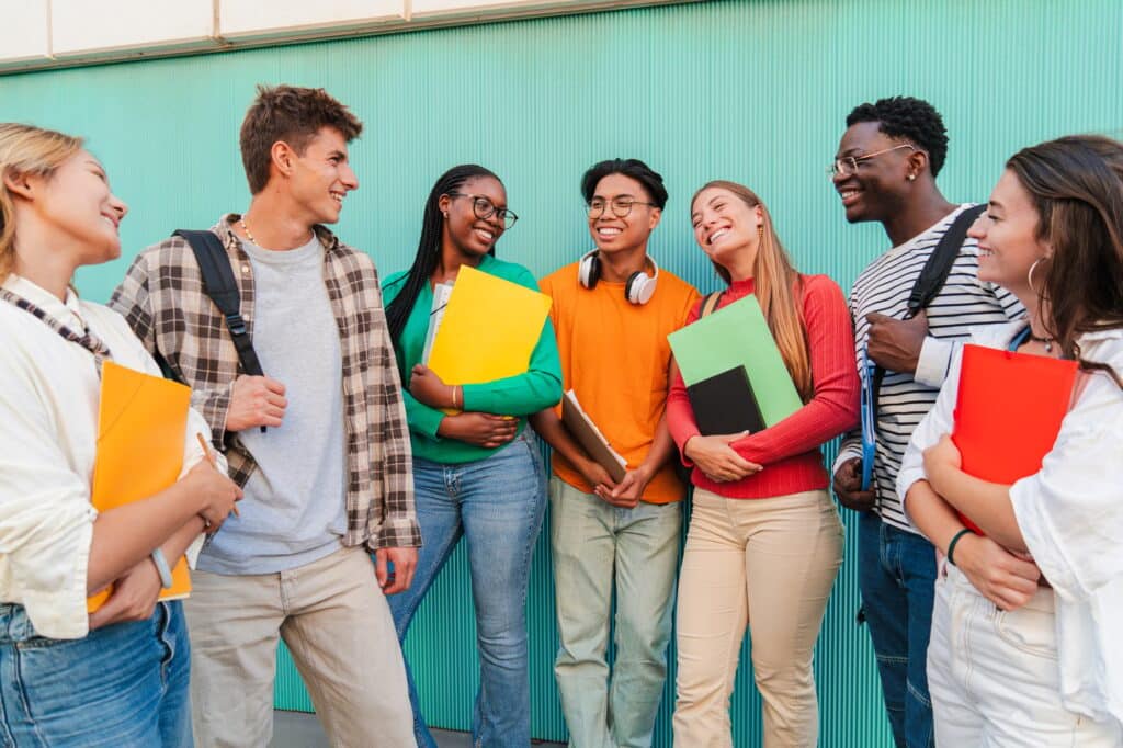 Diverse group of students talking and smiling outdoors, young friends holding notebooks and